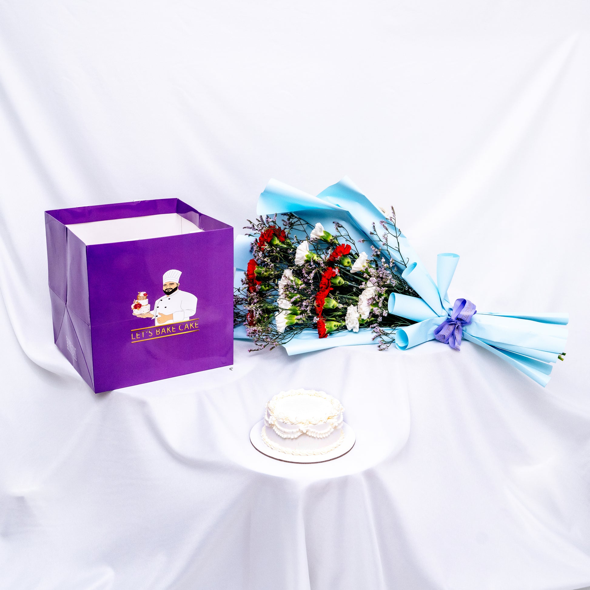 A premium gift set featuring a hand-piped white bento cake, a vibrant red and white flower bouquet in sky-blue wrap, and a signature 'Let's Bake Cake' purple gift bag on white silk, Abu Dhabi.