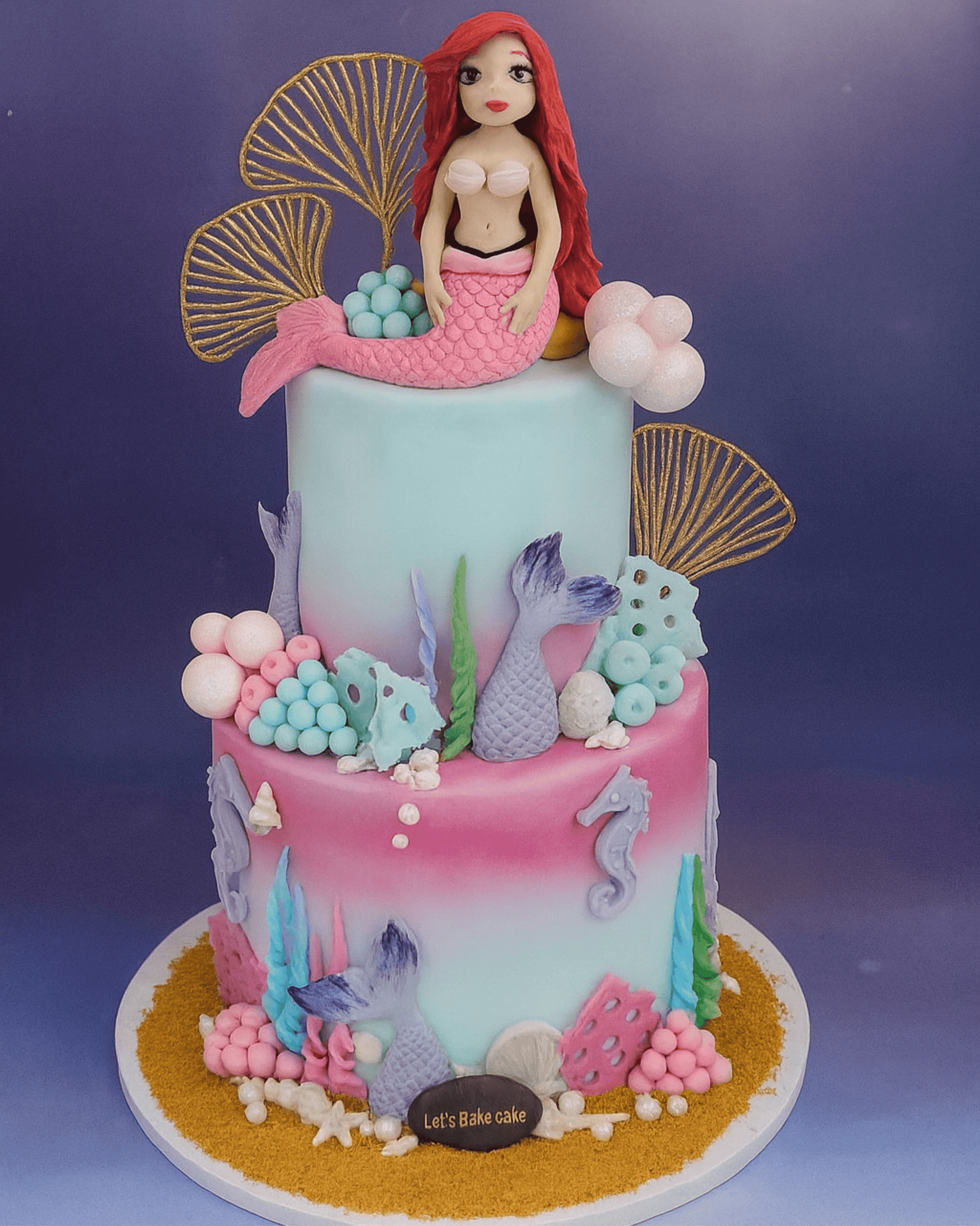 A luxury two-tiered Mermaid themed birthday cake with Ariel figurine, gold sea shells, and colorful coral decorations by Let's Bake Cake Abu Dhabi.