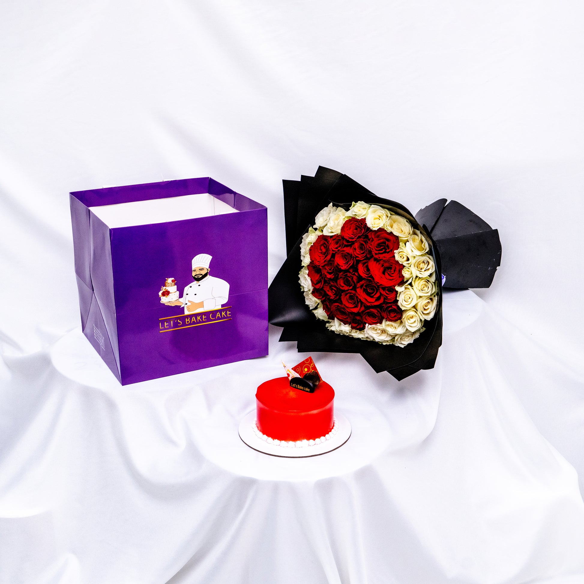 A luxury romantic gift set featuring a vibrant red glazed bento cake, a heart-shaped red and white rose bouquet in black premium wrap, and a branded 'Let's Bake Cake' purple gift bag on white silk, Abu Dhabi