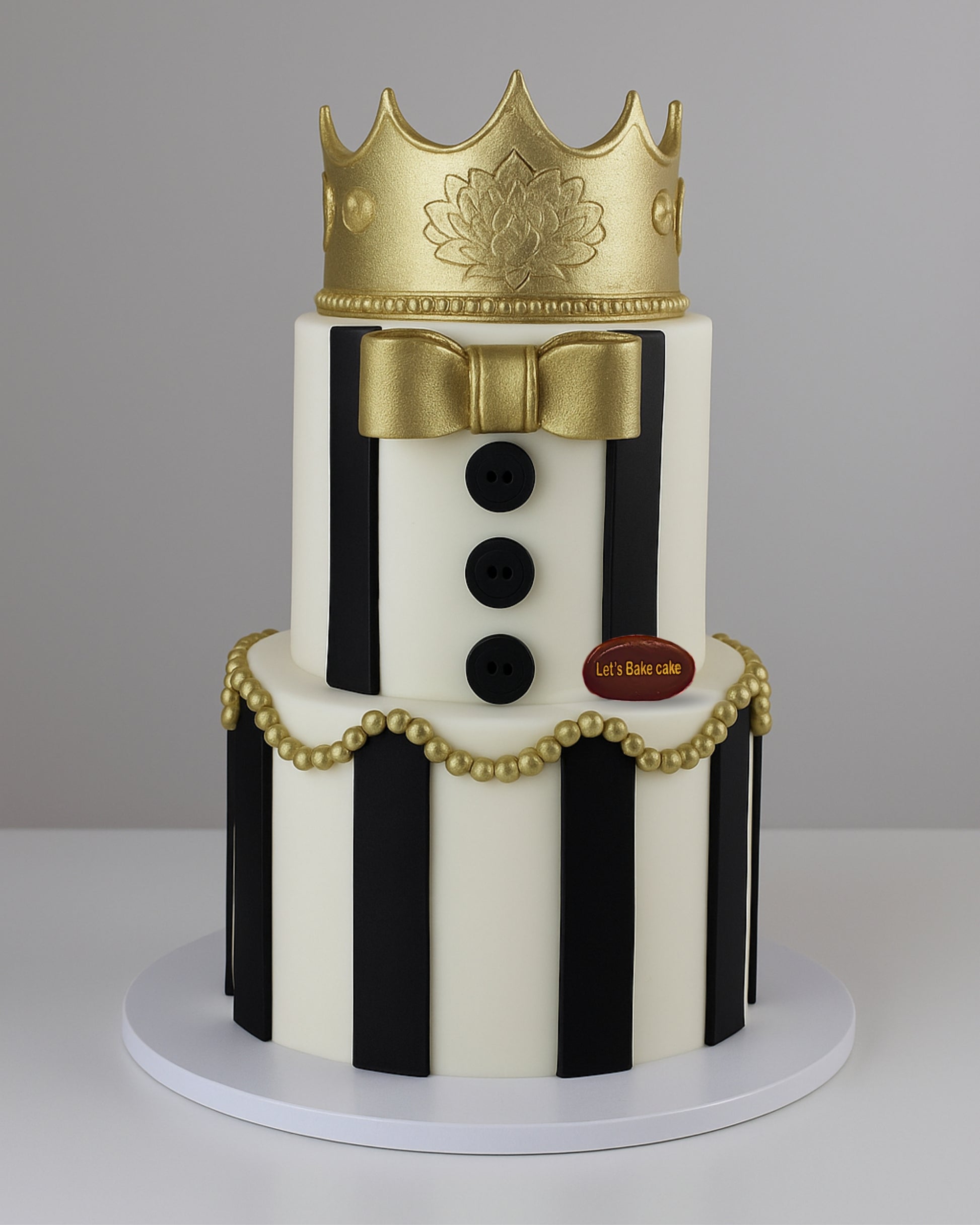 A luxury two-tiered black and white striped birthday cake topped with a majestic gold crown and gold pearl decorations by Let's Bake Cake Abu Dhabi.