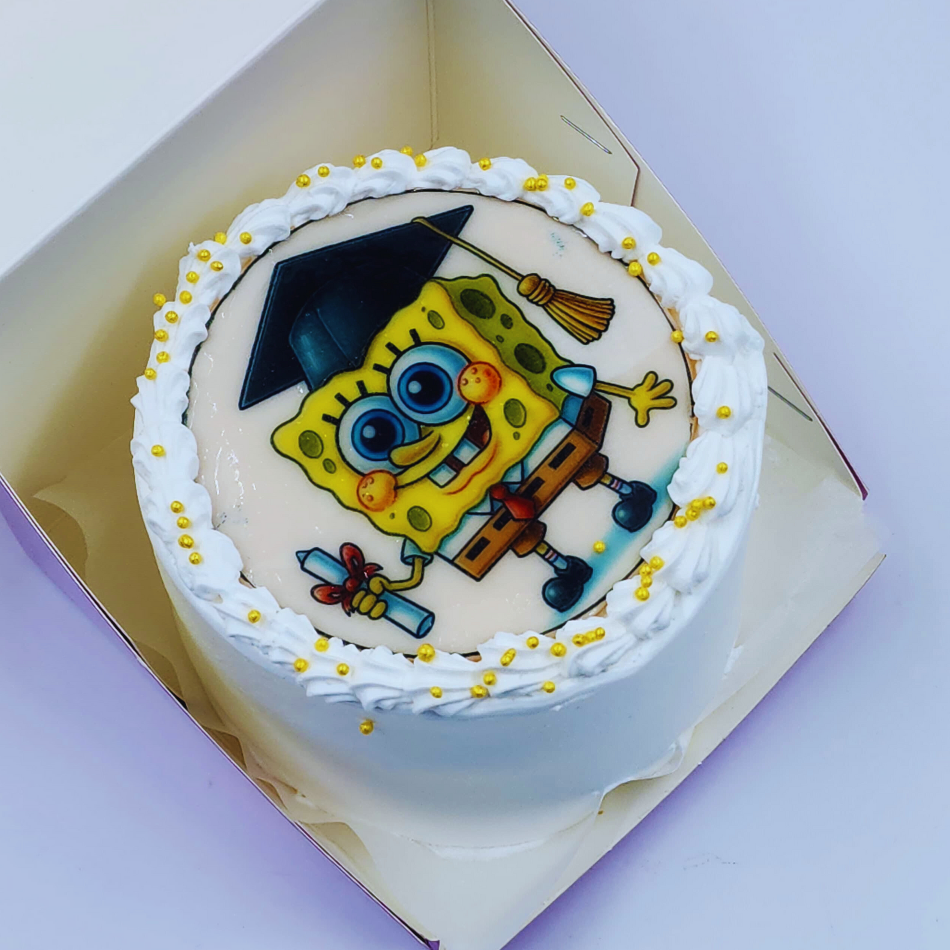A luxury white graduation cake with a SpongeBob SquarePants edible image and golden sprinkles by Let's Bake Cake Abu Dhabi.